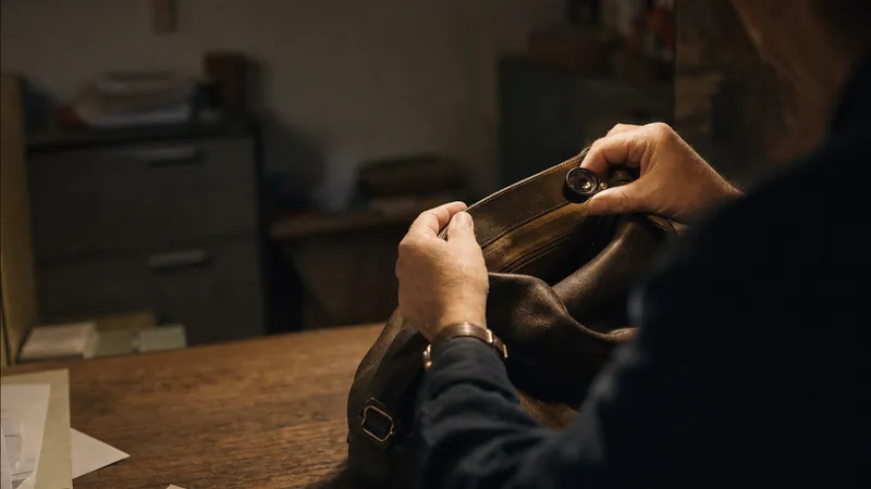 Hands holding a loupe close to the interior seam of an unbranded leather bag on a wooden counter