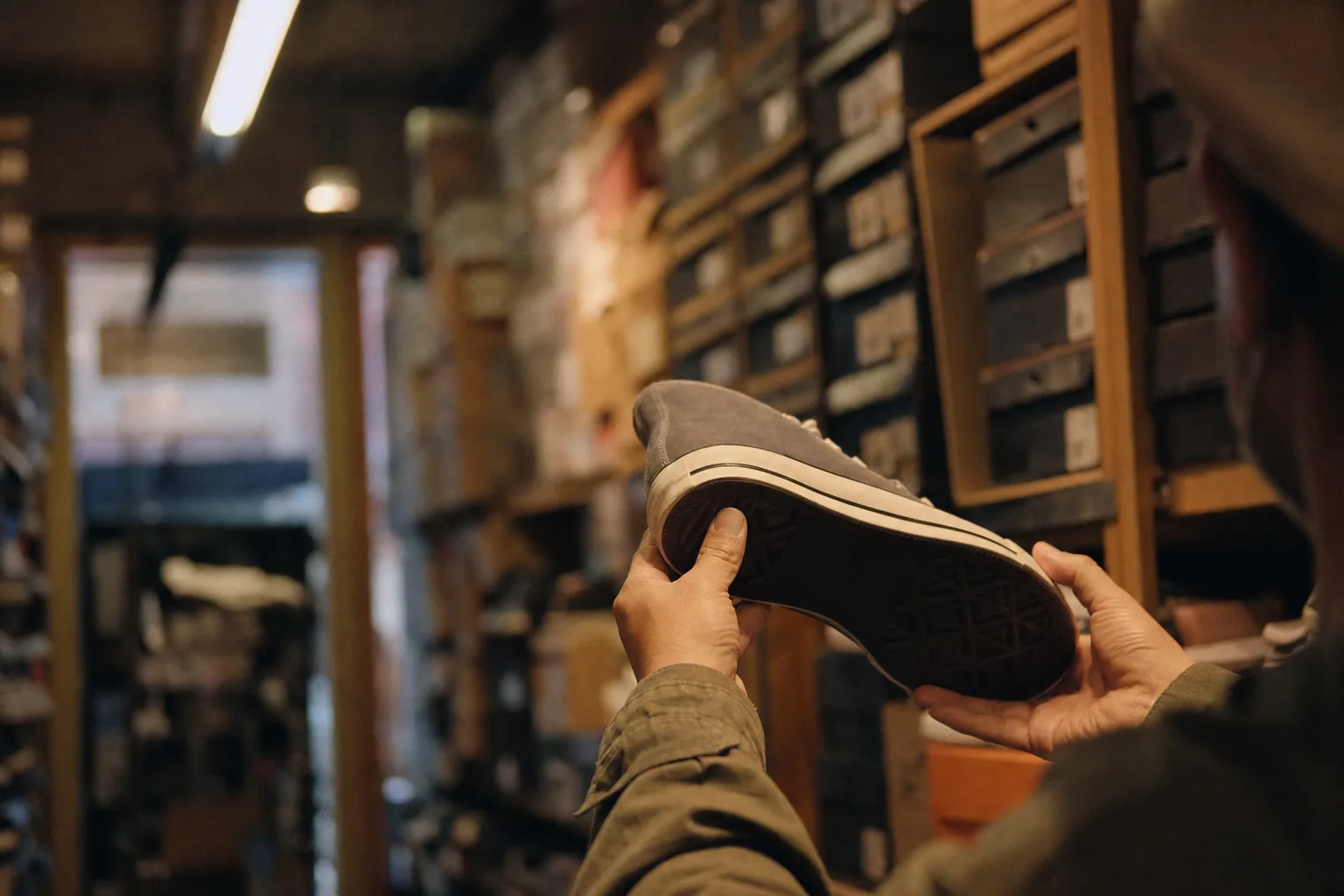 Hands inspecting the sole of an unbranded sneaker inside a narrow Japanese shoe shop with stacked boxes behind