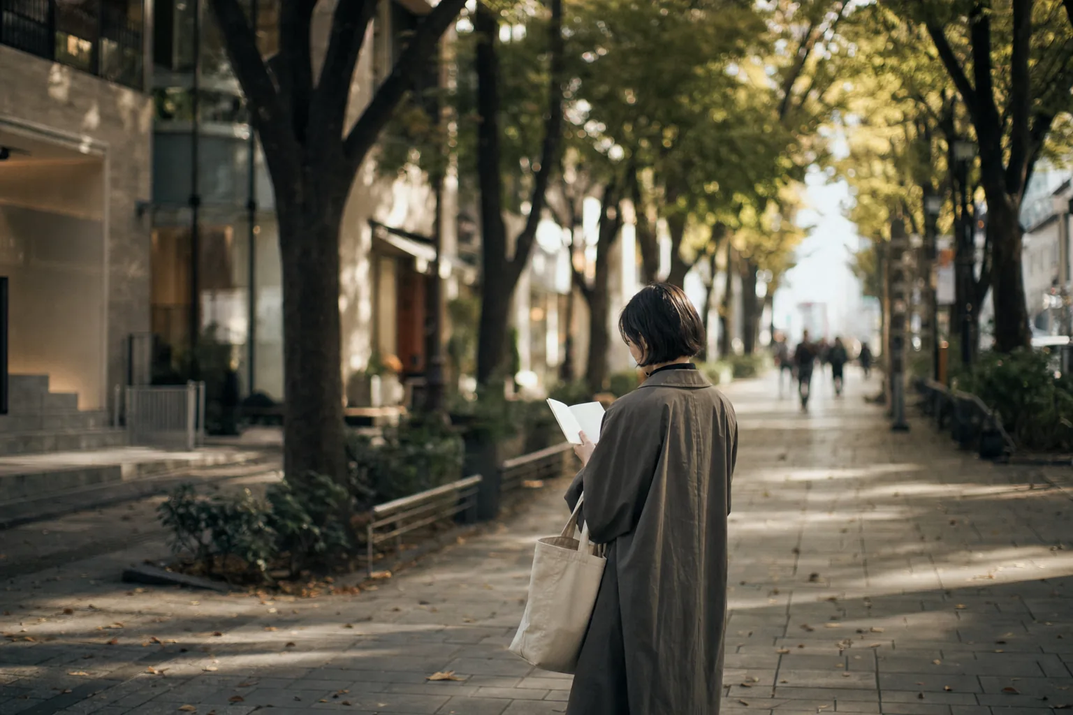 Shopper with notebook and tote bag standing on the tree-lined pavement of Omotesando Avenue, boutique facades blurred behind