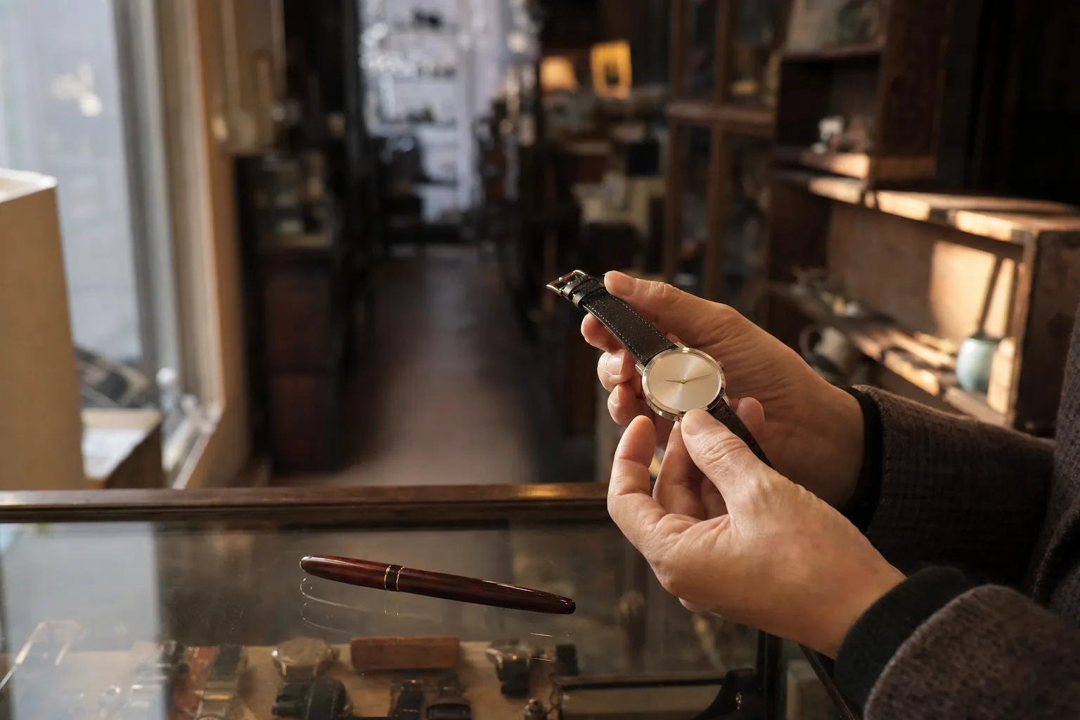 Hands examining an unbranded silver dress watch beside a deep-red lacquer fountain pen on a glass shop counter
