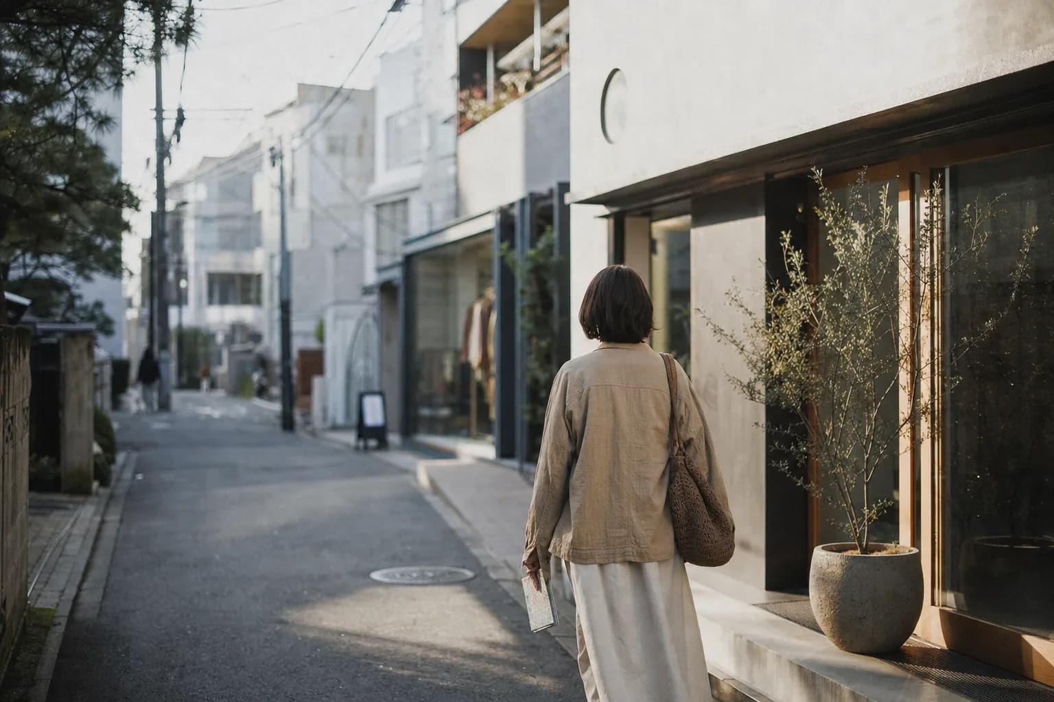 A lone shopper walks a narrow Tokyo boutique side street, notebook in hand, in soft morning light