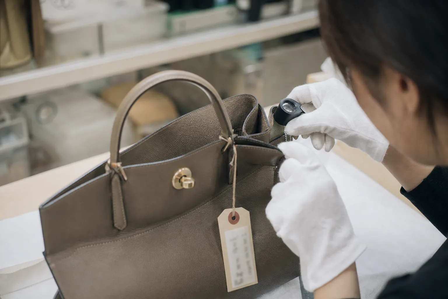 Hands holding a loupe over the corner of an unbranded leather handbag on a white cloth, with a blurred Japanese condition